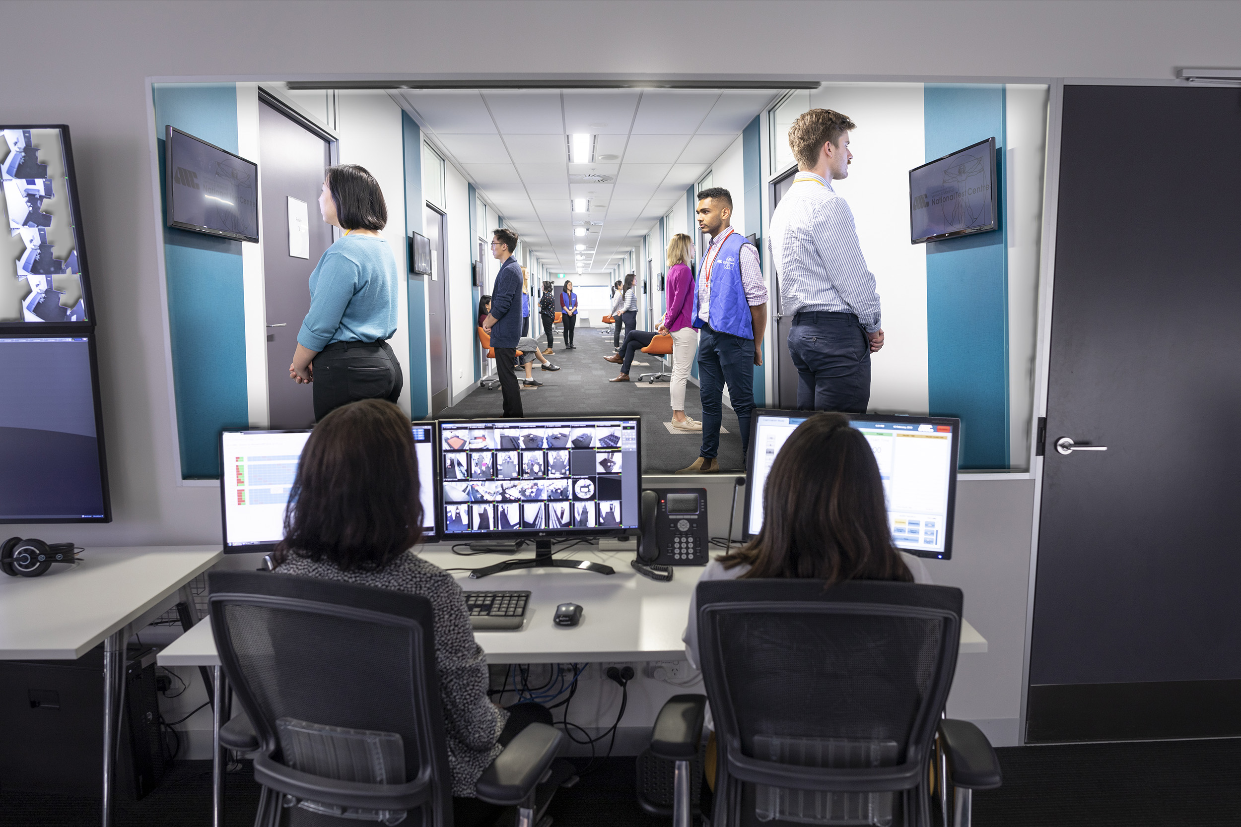 two women sitting in a control room looking at computer screens. In front of them is a window which shows a view into a corridor where people are standing in front of doors waiting to go in.