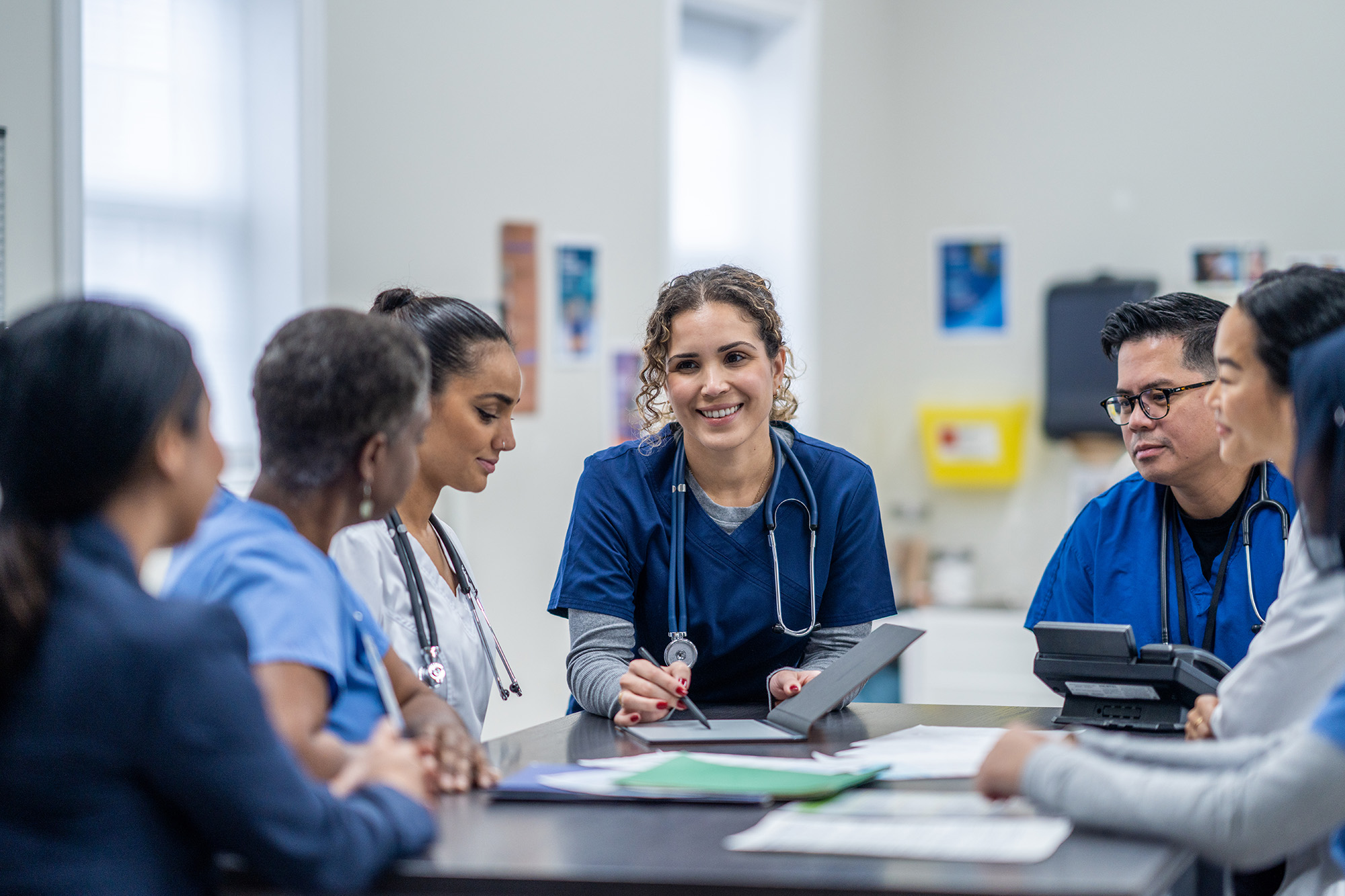 A group of healthcare professionals in scrubs and lab coats sit around a table in a meeting room, discussing documents and smiling. One person in the center is holding a tablet.