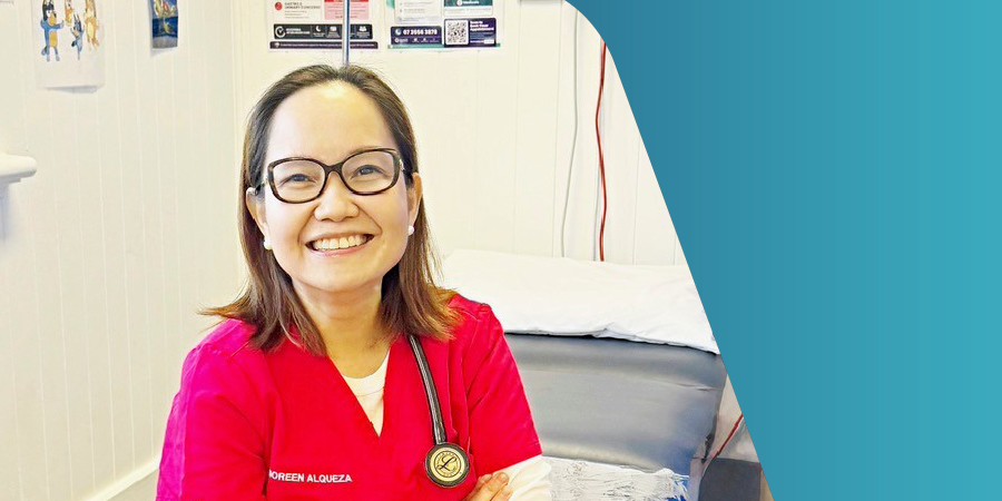A doctor sitting on a bed in a doctor's surgery, wearing red scrubs with a stethoscope around her neck smiling at the camera